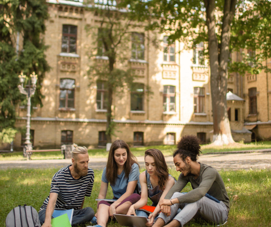 Four students discuss a topic outside a college building, seated on a grassy area.